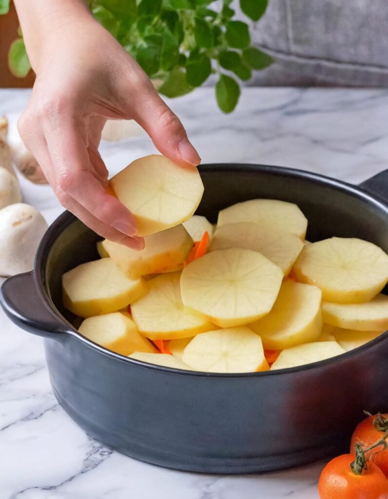 placing potato In a kitchen with a white marble countertop, a black crockpot is the central focus. A human hand is placing thinly sliced rounds of orange sweet potatoes on top of white potatoes already layered inside the crockpot. The potatoes are arranged in an overlapping pattern, covering the bottom of the crockpot. The hand appears to be in the middle of the layering process, suggesting the preparation of a dish that will be slow-cooked to perfection. The scene gives an impression of home cooking and the preparation of a comforting meal. There is a blue and white checkered cloth partially visible in the background, adding a touch of homely decor to the setting.